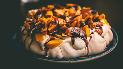 Photo of a pavlova indulgently decorated with chopped fruit and sauce. 