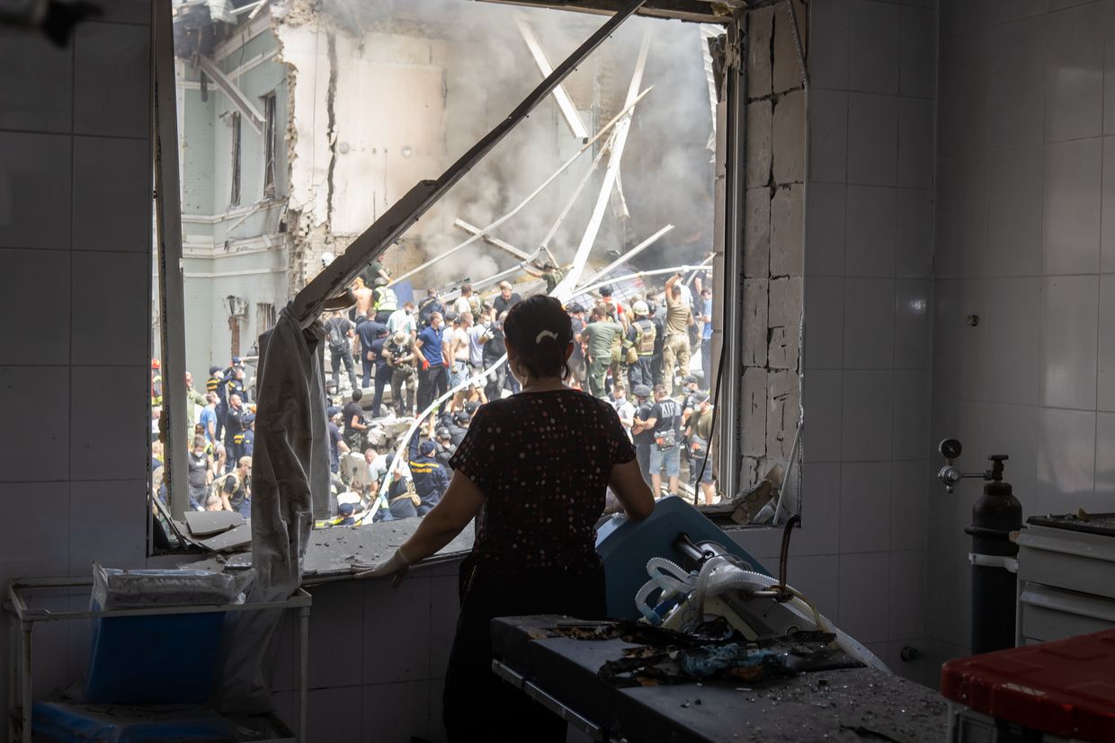 A healthcare worker looks through a window of a damaged operating room at people clear rubble at the building of one of the largest children's hospitals of Ukraine, Okhmatdyt